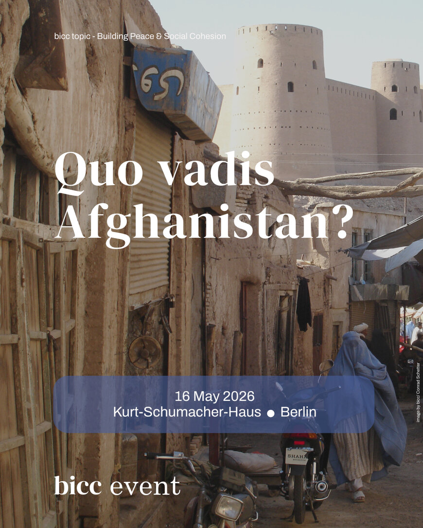 Narrow street in an old town with mud-brick buildings and a person in traditional clothing walking past a motorcycle with a fortress wall in the background